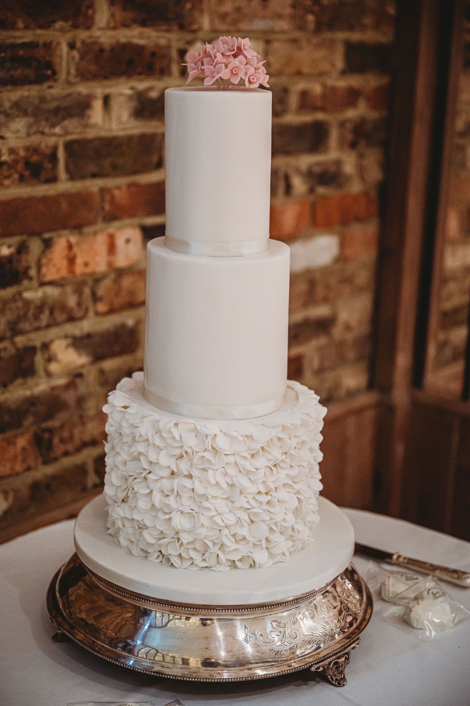 Elegant three-tier white wedding cake with ruffled fondant bottom layer and blush pink flowers on top, displayed on silver stand.