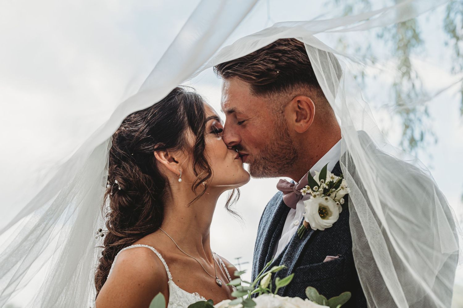 A romantic embracing couple in wedding attire share a kiss under a flowing white veil on their special day.
