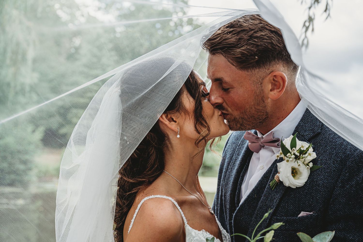 A bride and groom share a romantic kiss under a flowing wedding veil in an outdoor setting.