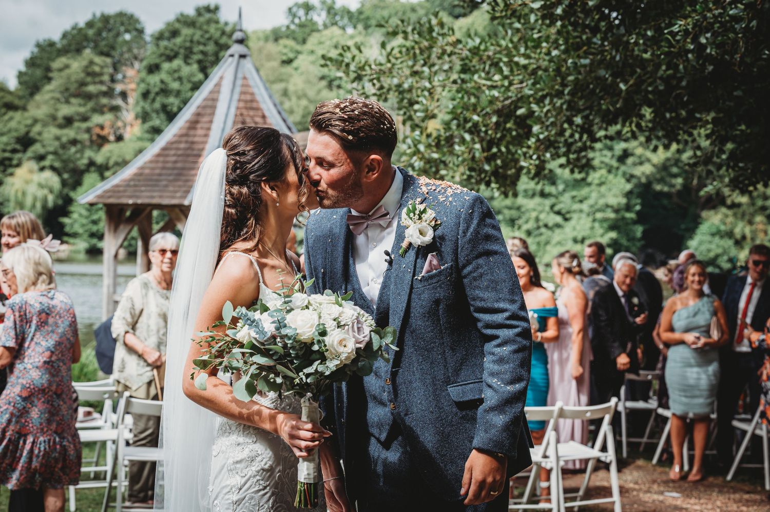 A couple shares an intimate moment at an outdoor wedding ceremony by a gazebo surrounded by guests and greenery.