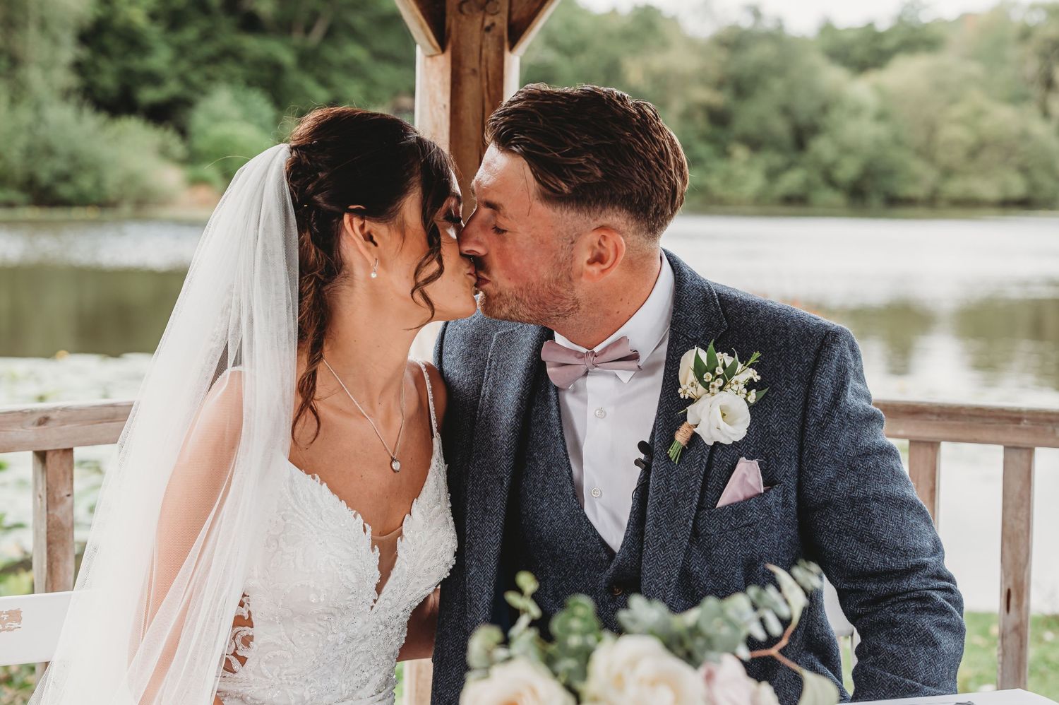 A romantic wedding couple sharing a kiss at a lakeside gazebo, with a white boutonniere and veil visible.