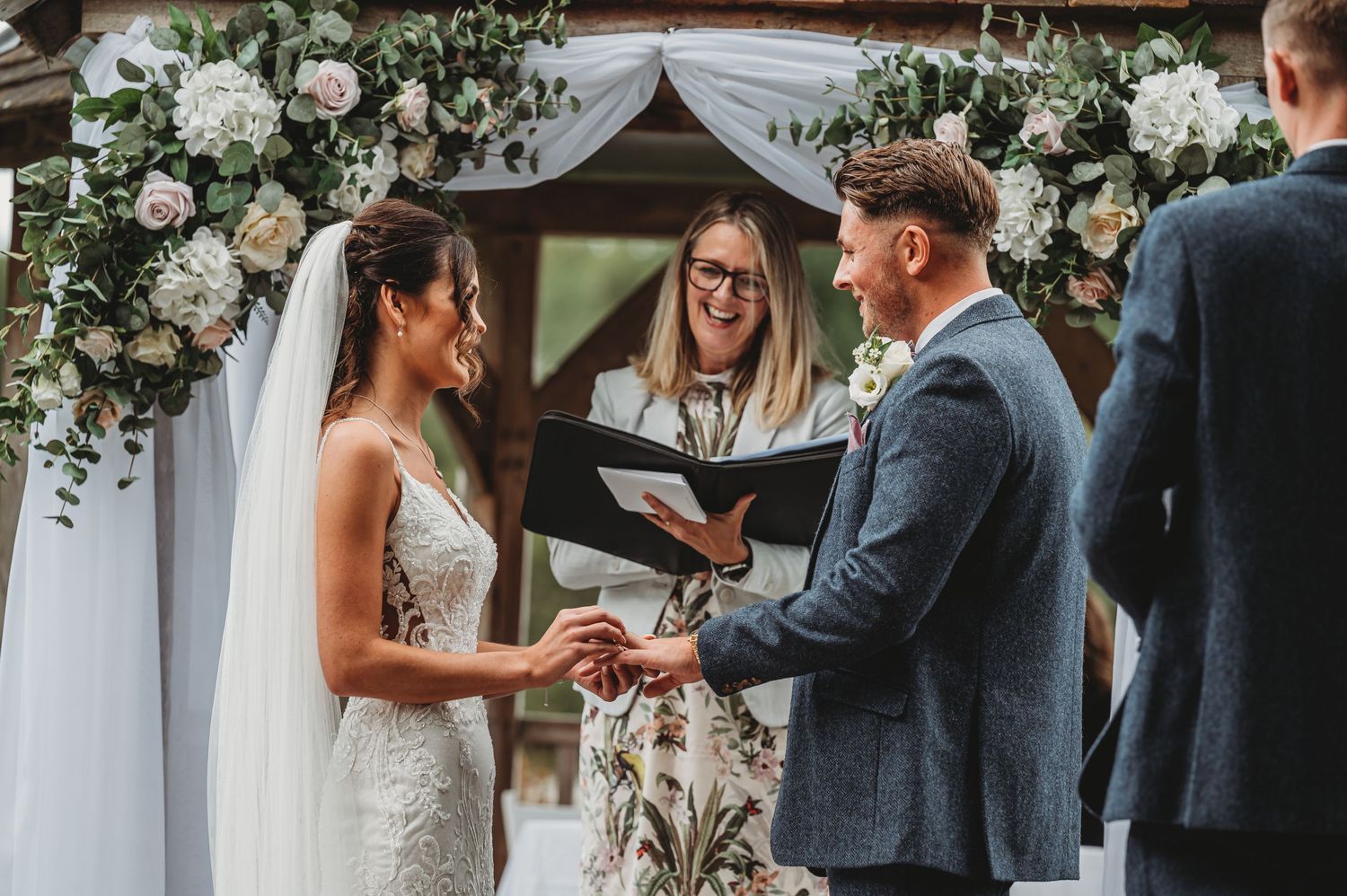 A wedding ceremony takes place under a beautiful white floral arch with an officiant presiding over the exchange of rings.