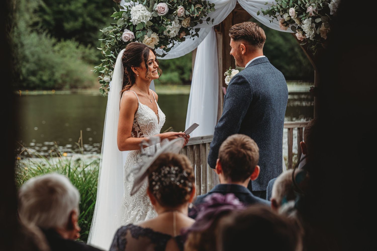 A couple exchanges vows during an outdoor lakeside wedding ceremony with guests watching beneath a floral arch.