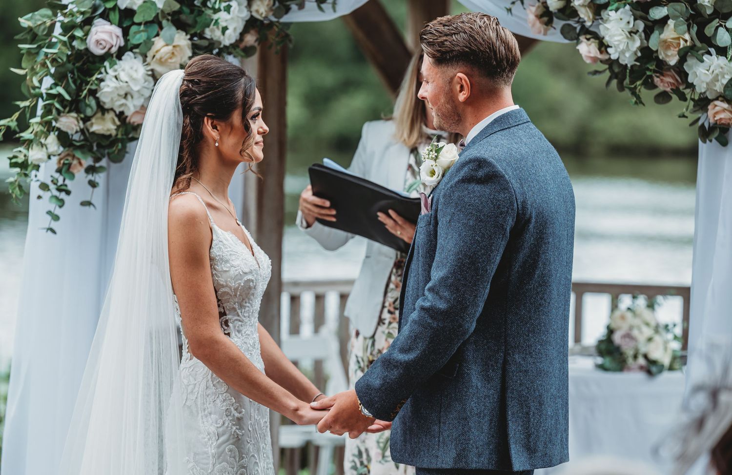 Couple exchanges vows under floral arch during outdoor waterfront wedding ceremony in natural setting.