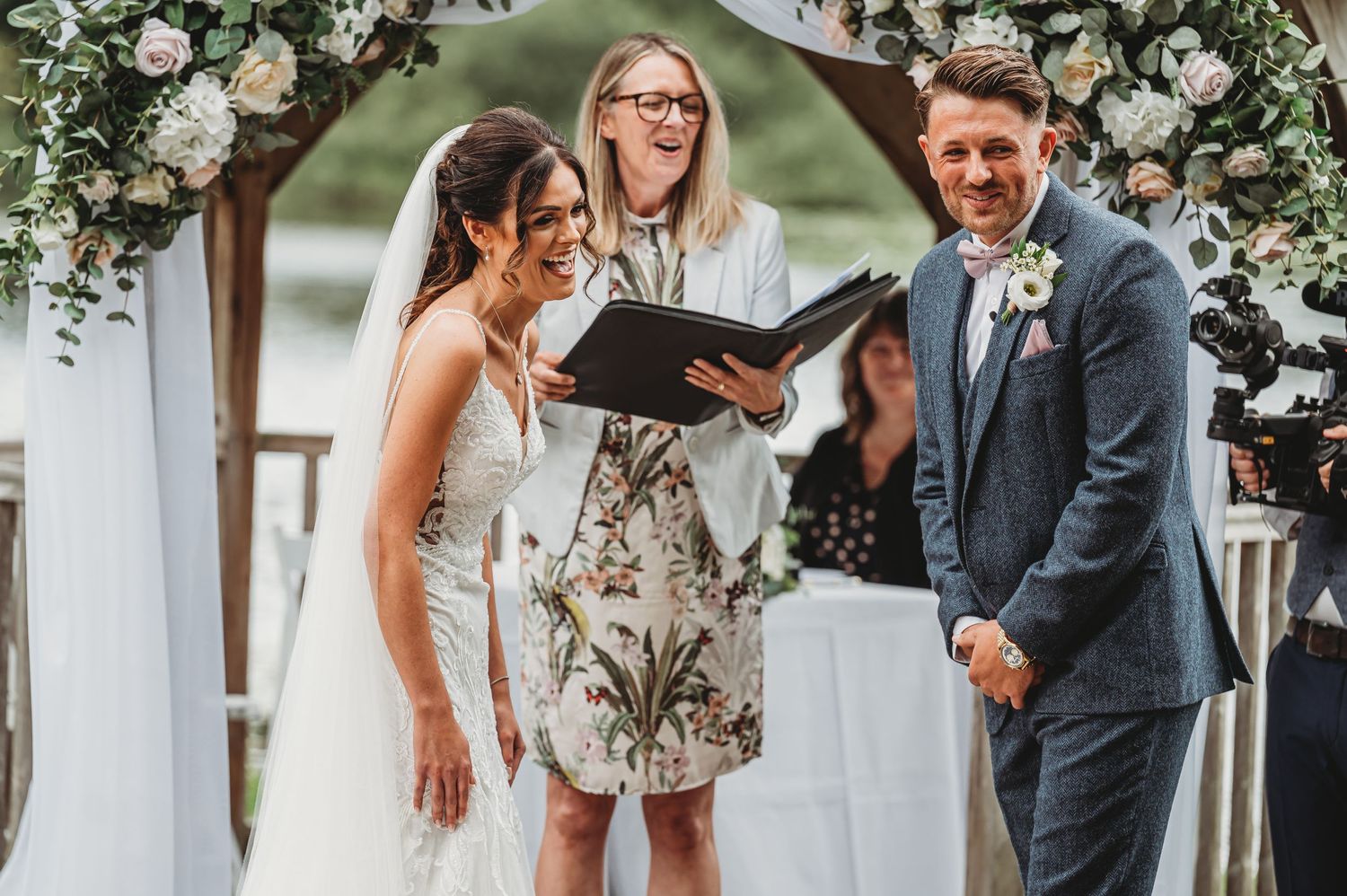 A joyous wedding ceremony takes place under a floral arch with white roses as an officiant reads from a book.