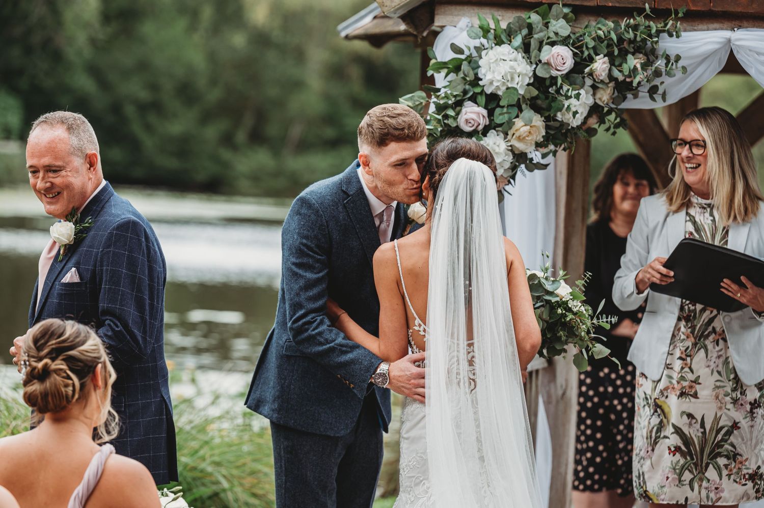 A romantic outdoor wedding ceremony takes place by a lake with white floral decorations and ceremony arch.