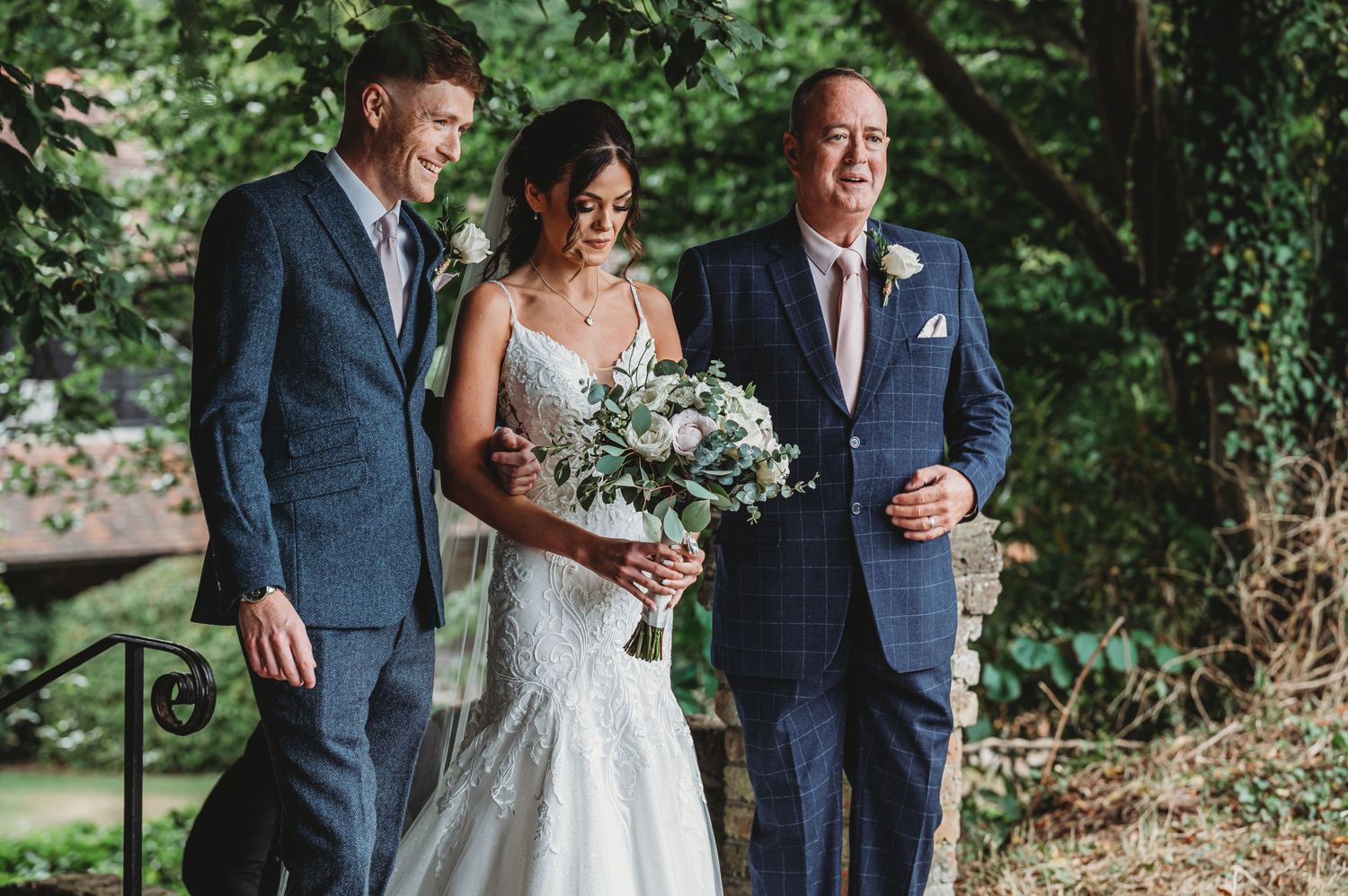 A joyful outdoor wedding moment captured in nature with three people in formal attire and a white floral bouquet.