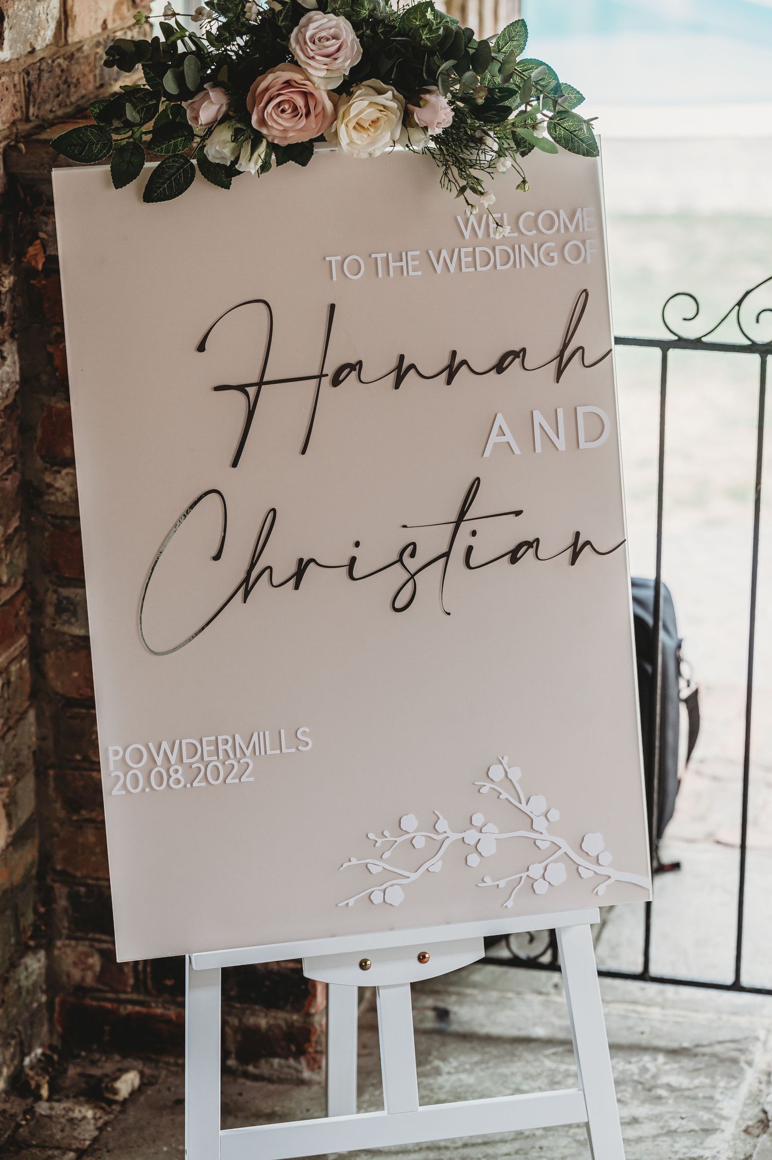 Elegant wedding welcome sign with blush roses and greenery displays couple names in modern calligraphy script.