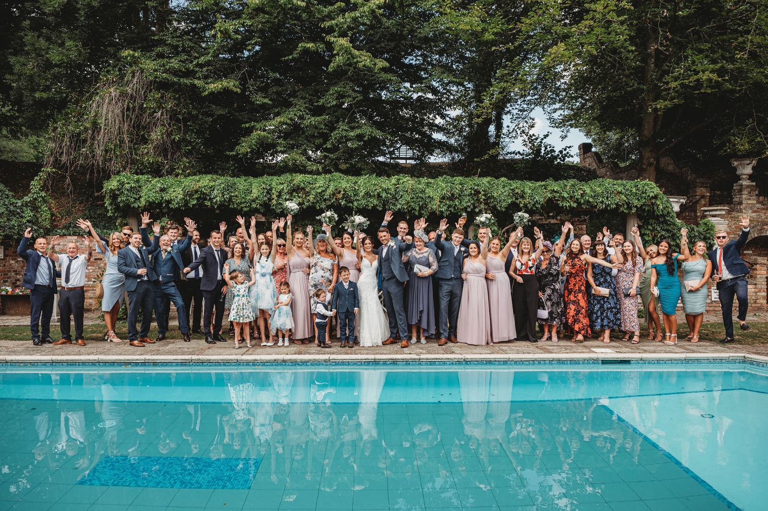 Wedding party and guests gather for a group photo beside a bright blue swimming pool surrounded by trees.