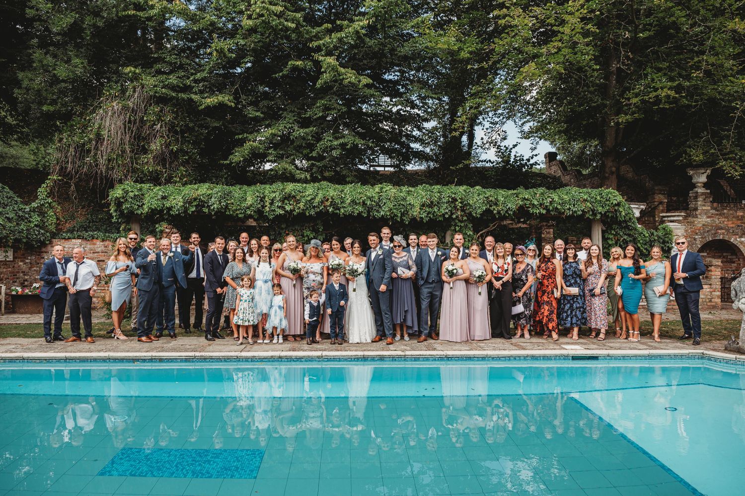 Large wedding party gathered beside outdoor swimming pool at elegant venue for group photo.