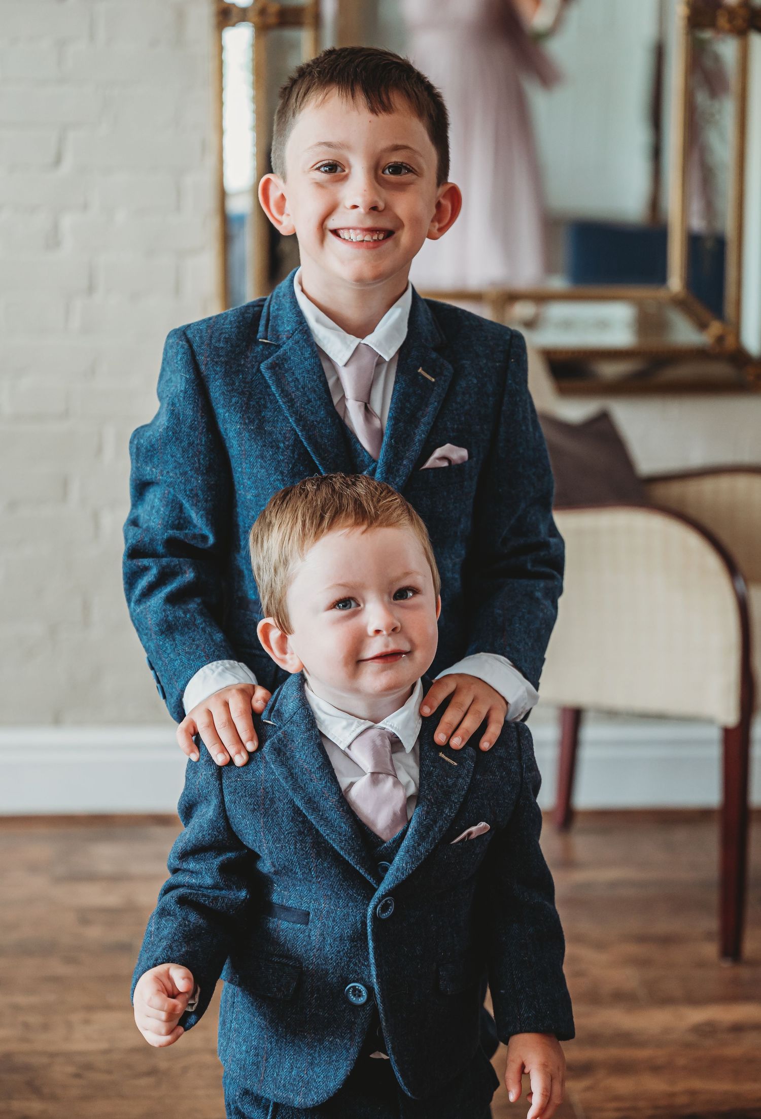 Two young boys wearing matching blue suits and pink ties smile for a formal portrait on a hardwood floor.