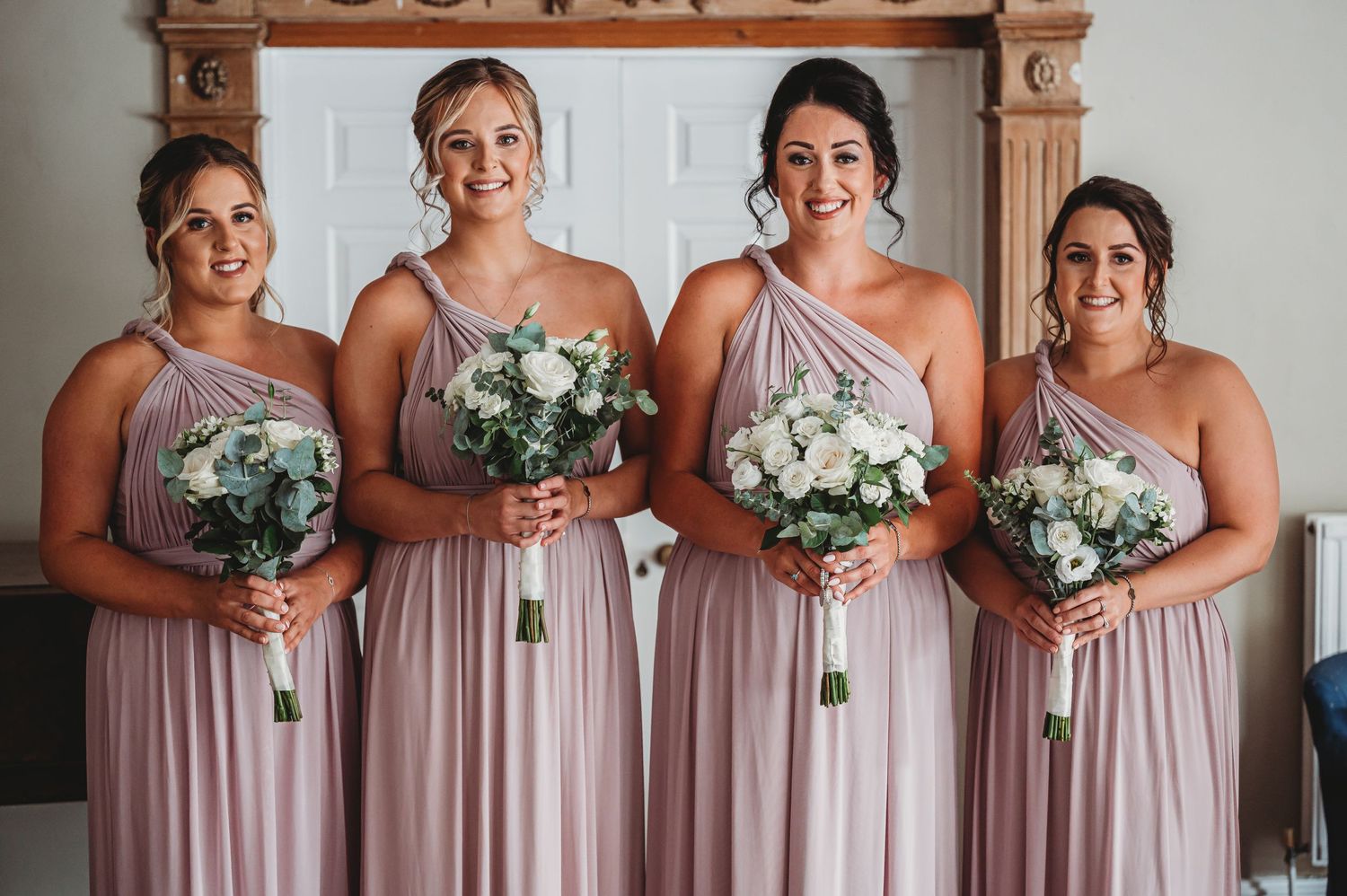 Bridesmaids wearing blush pink one-shoulder dresses pose together while holding white rose bouquets.