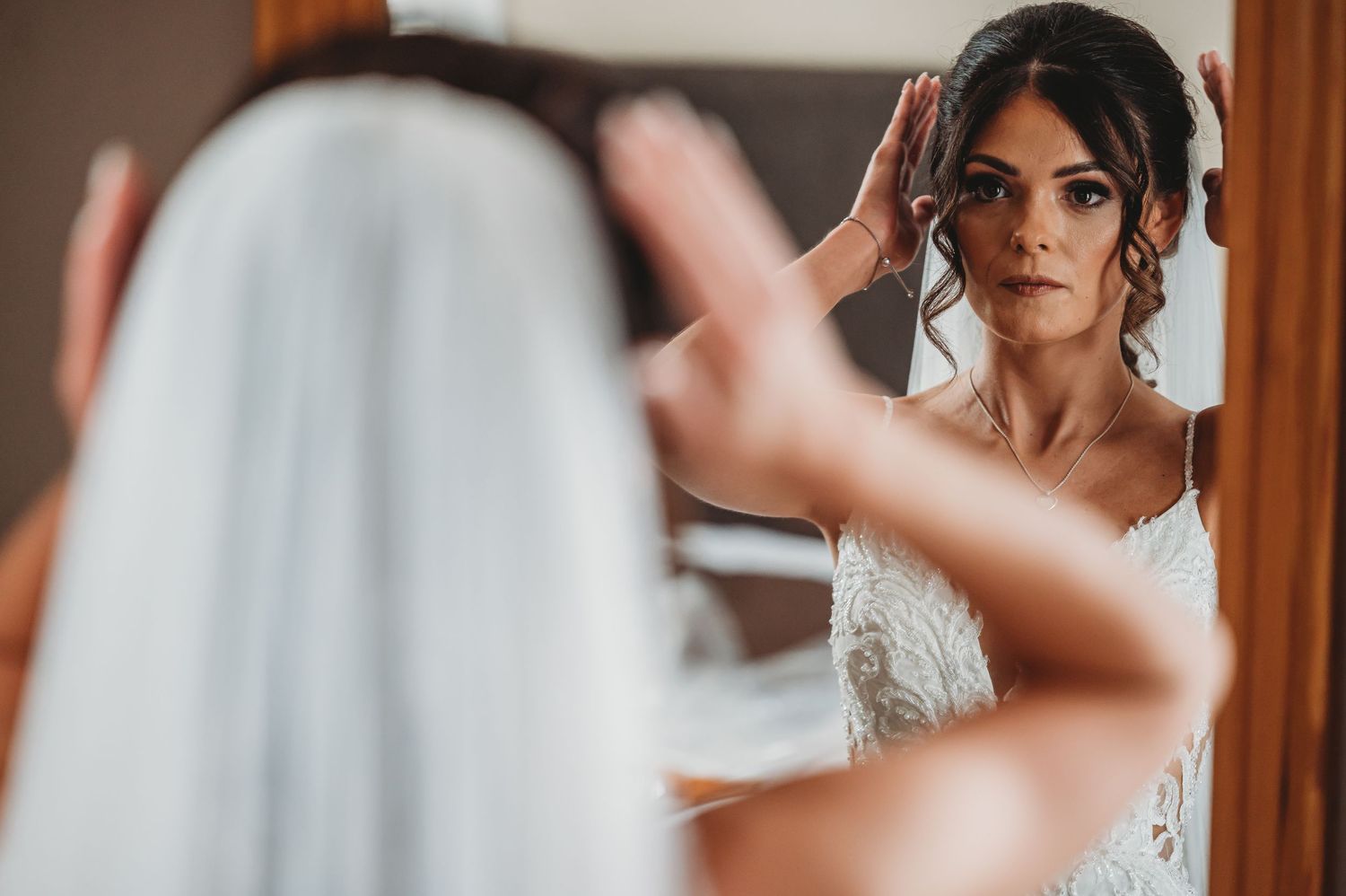 A bride in a white lace wedding dress looks at her reflection in a mirror while getting ready.