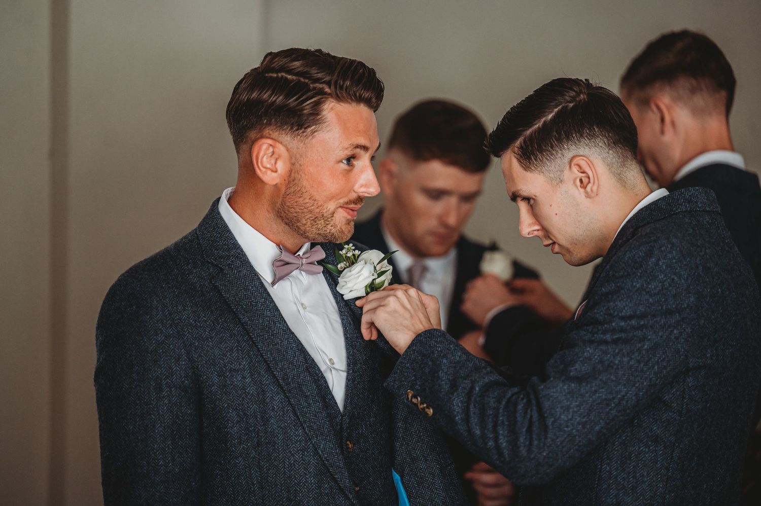 Groomsmen helping to adjust boutonniere on navy suit with bow tie during wedding preparations.