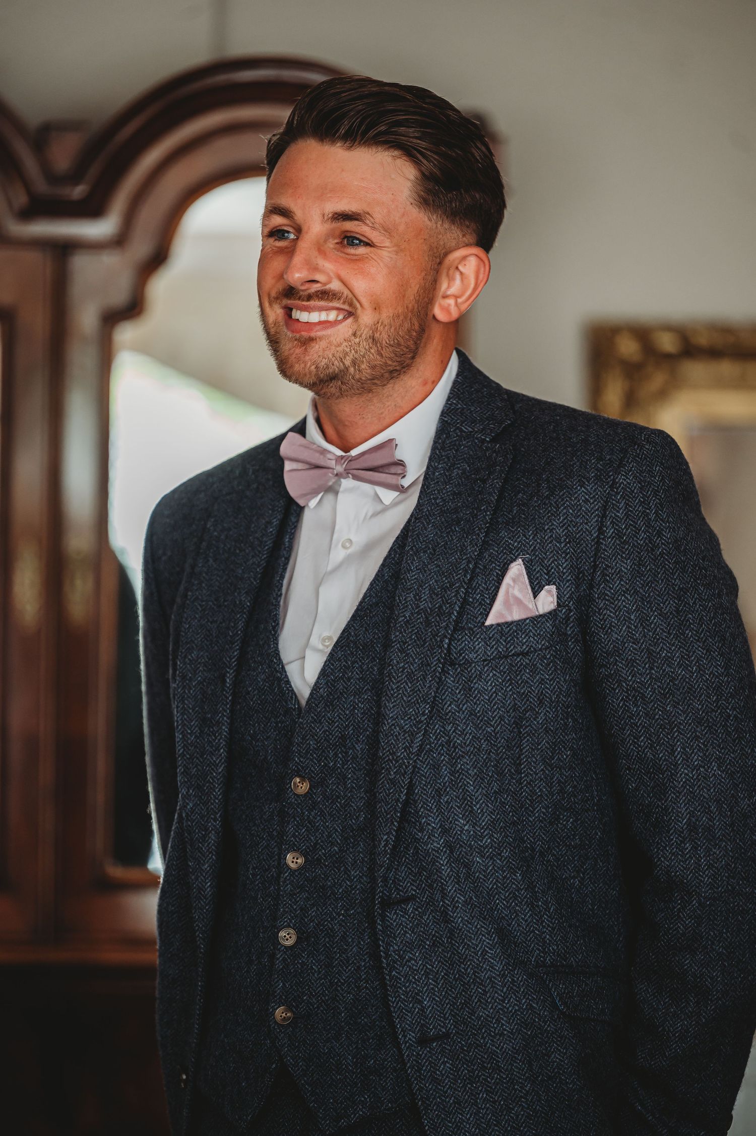 A groom in a charcoal three-piece suit with pink bow tie and pocket square smiles warmly on his wedding day.