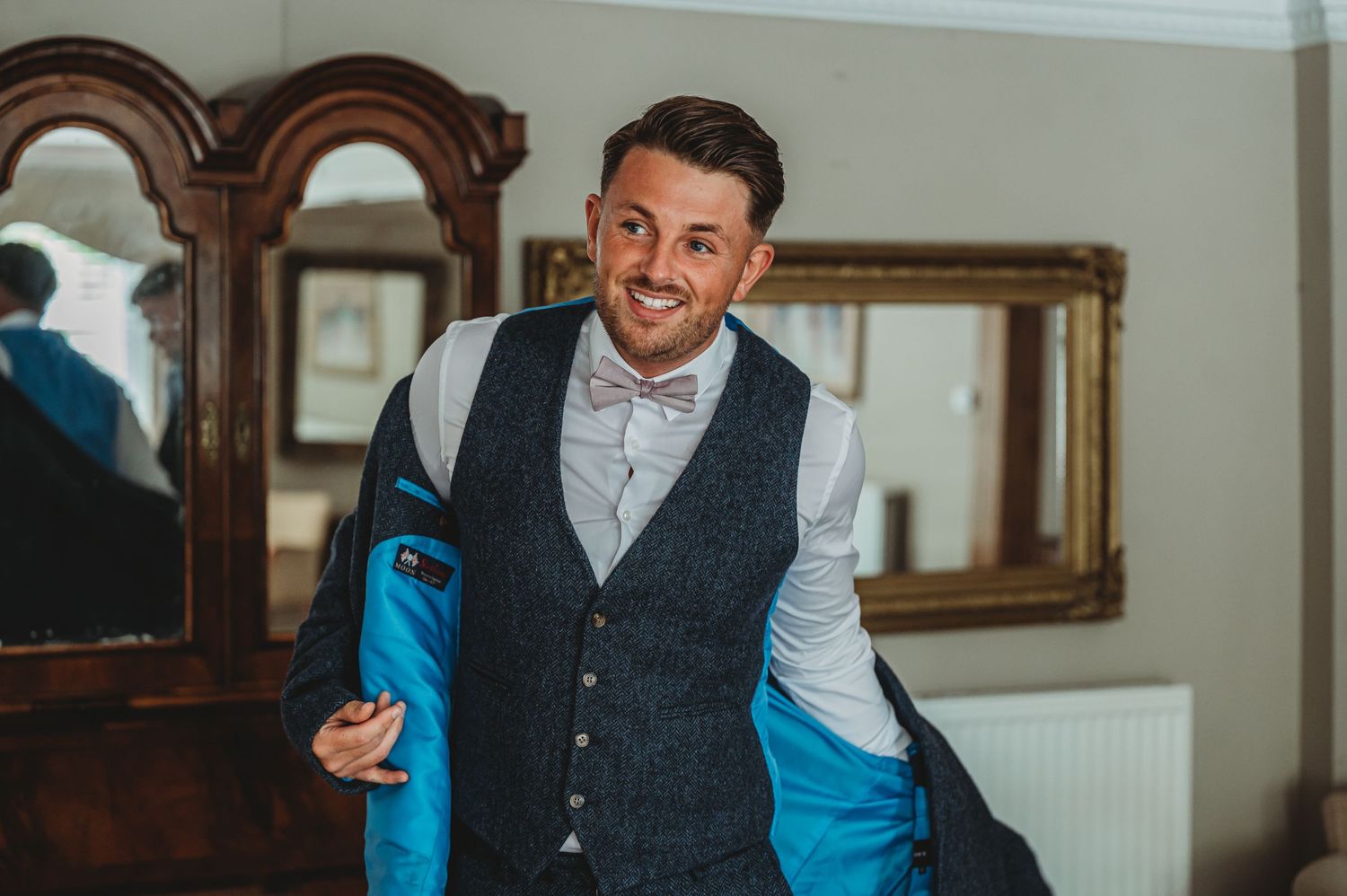 A person in a gray waistcoat and bowtie smiles while standing in a room with ornate mirrors.