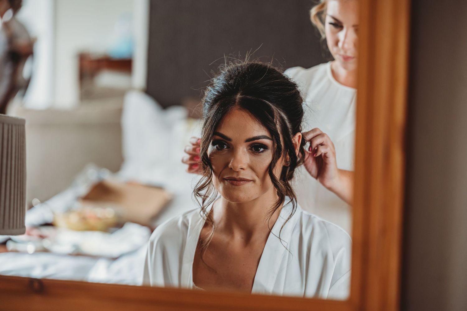 Wedding preparations scene with stylist arranging bride's updo hairstyle in warm morning light.