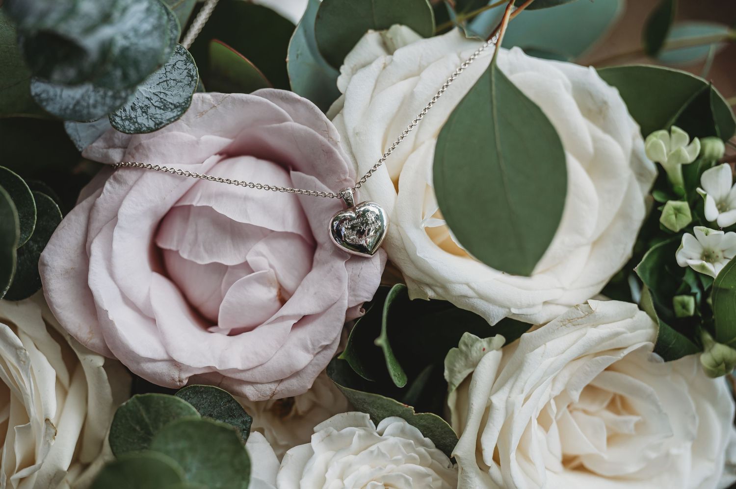 A delicate heart pendant necklace rests among soft pink and white roses with green foliage.