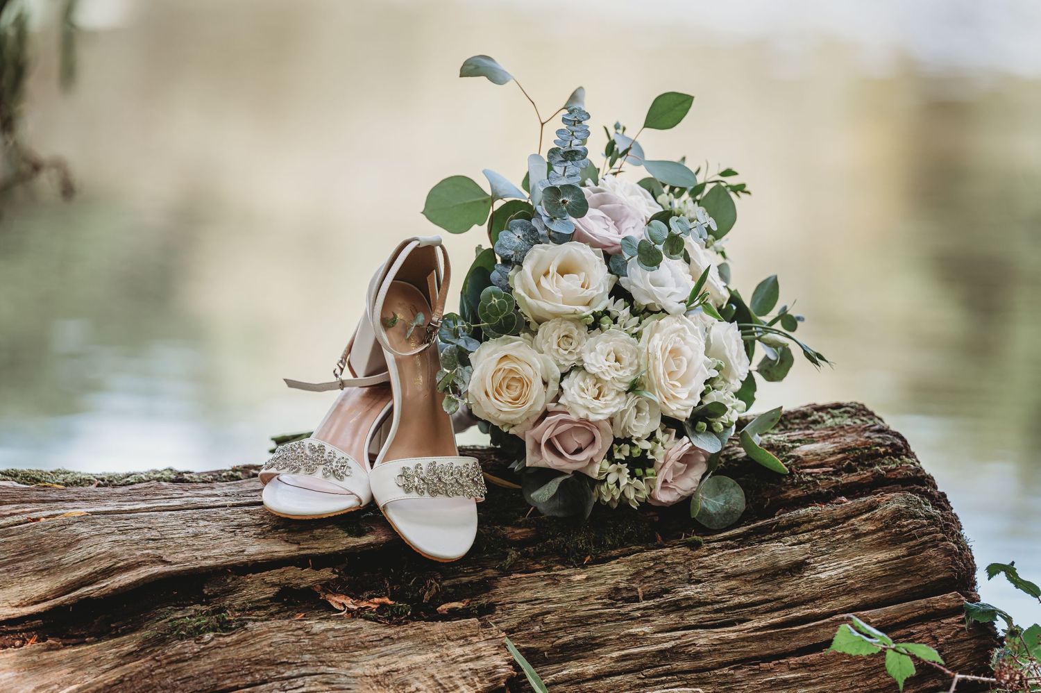 Elegant bridal shoes and white rose bouquet arranged on rustic driftwood by waterfront.