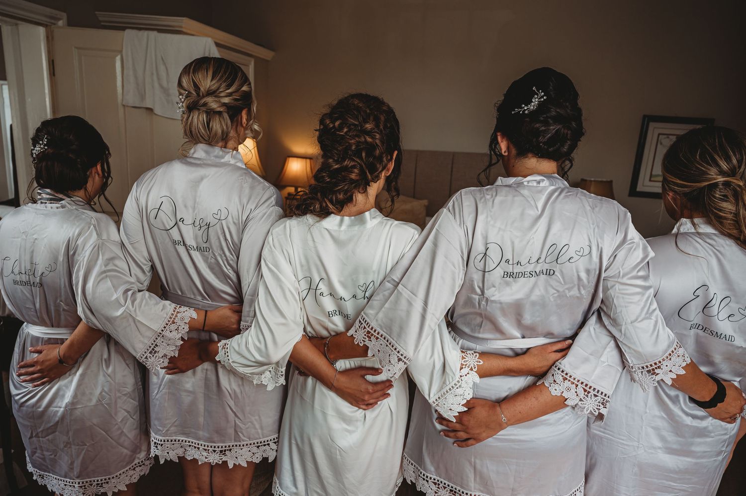 A group in matching white silk robes stands with their backs to the camera in a bedroom setting.
