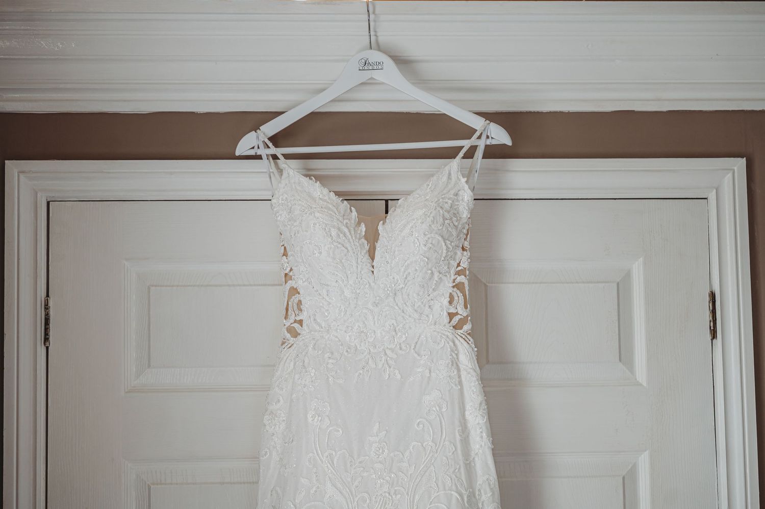 A delicate white lace wedding dress with spaghetti straps hangs from a white hanger against a white door.