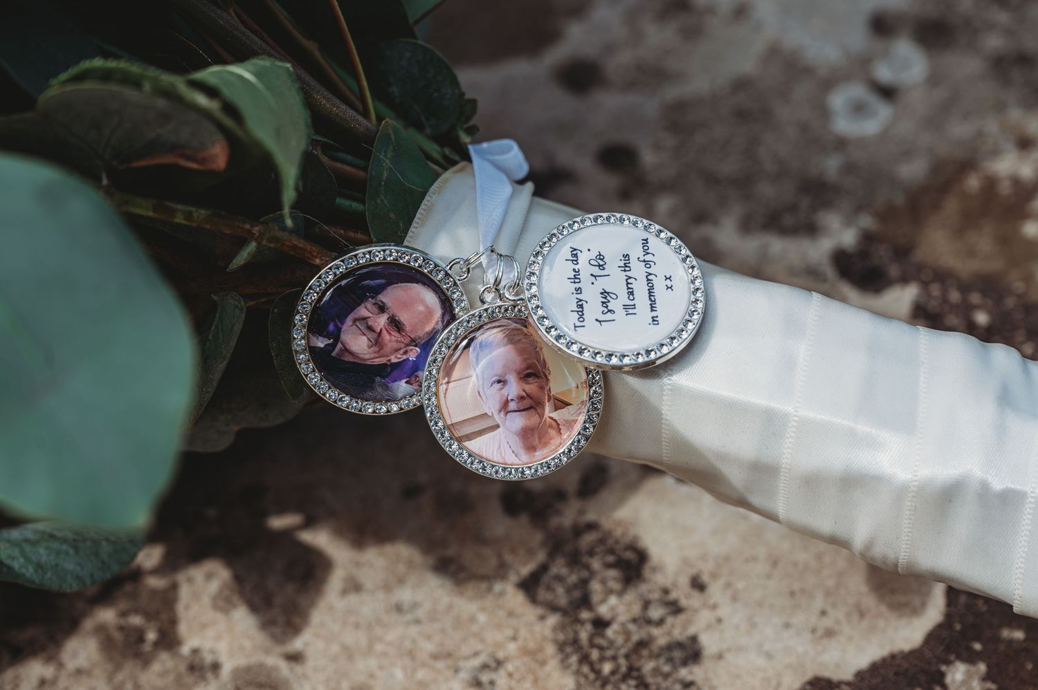 A wedding bouquet detail shows two circular photo charm pendants attached to white ribbon with greenery in the background.