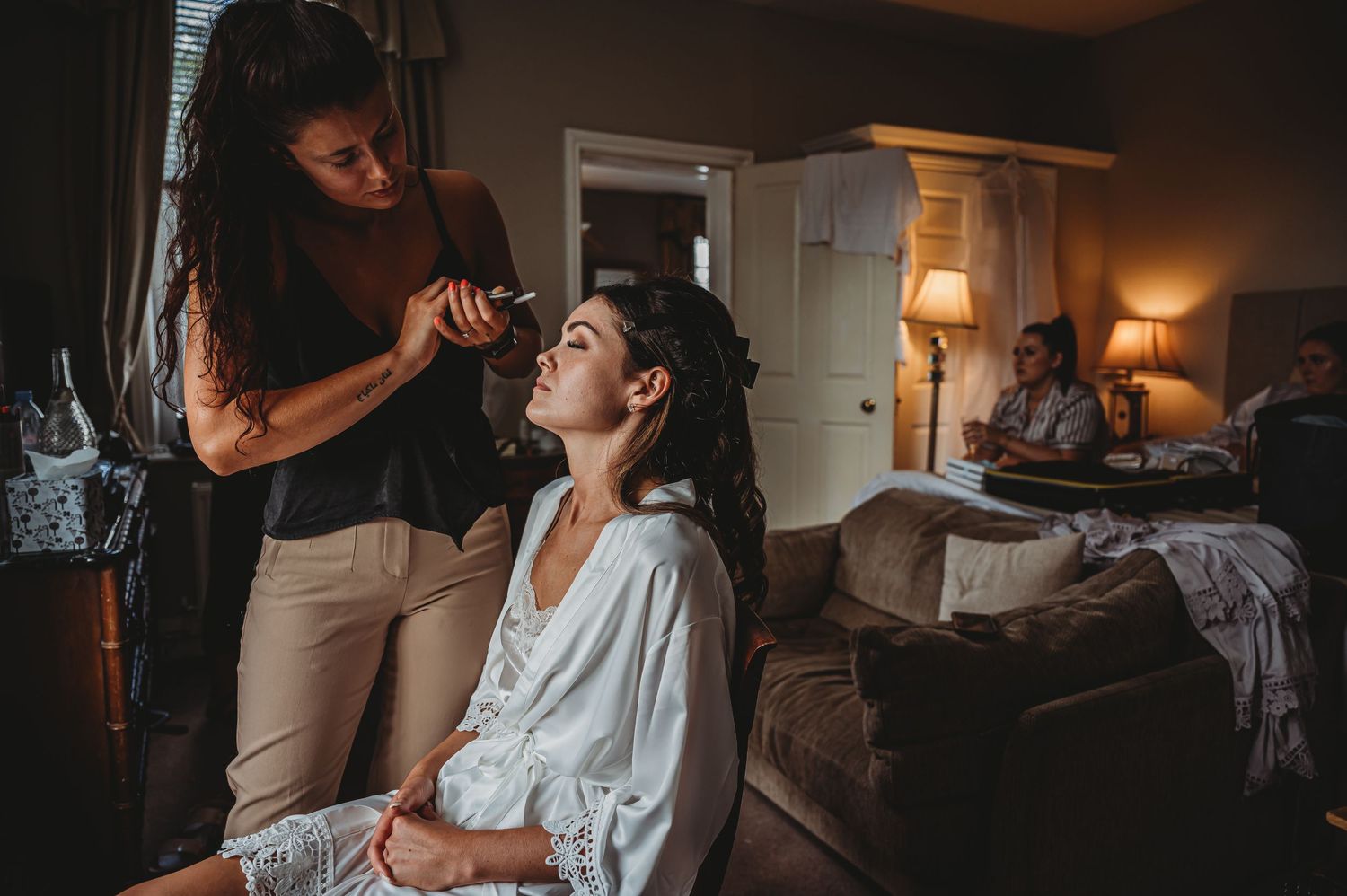 Makeup artist applies cosmetics to client in dim-lit room with warm lamps and cozy interior setting.