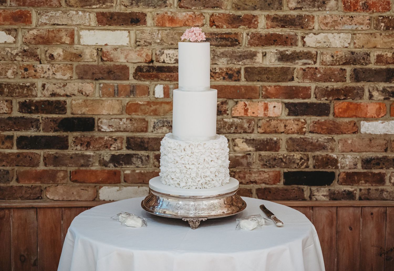 White five-tier wedding cake with delicate lace details sits on silver stand against rustic brick wall backdrop.