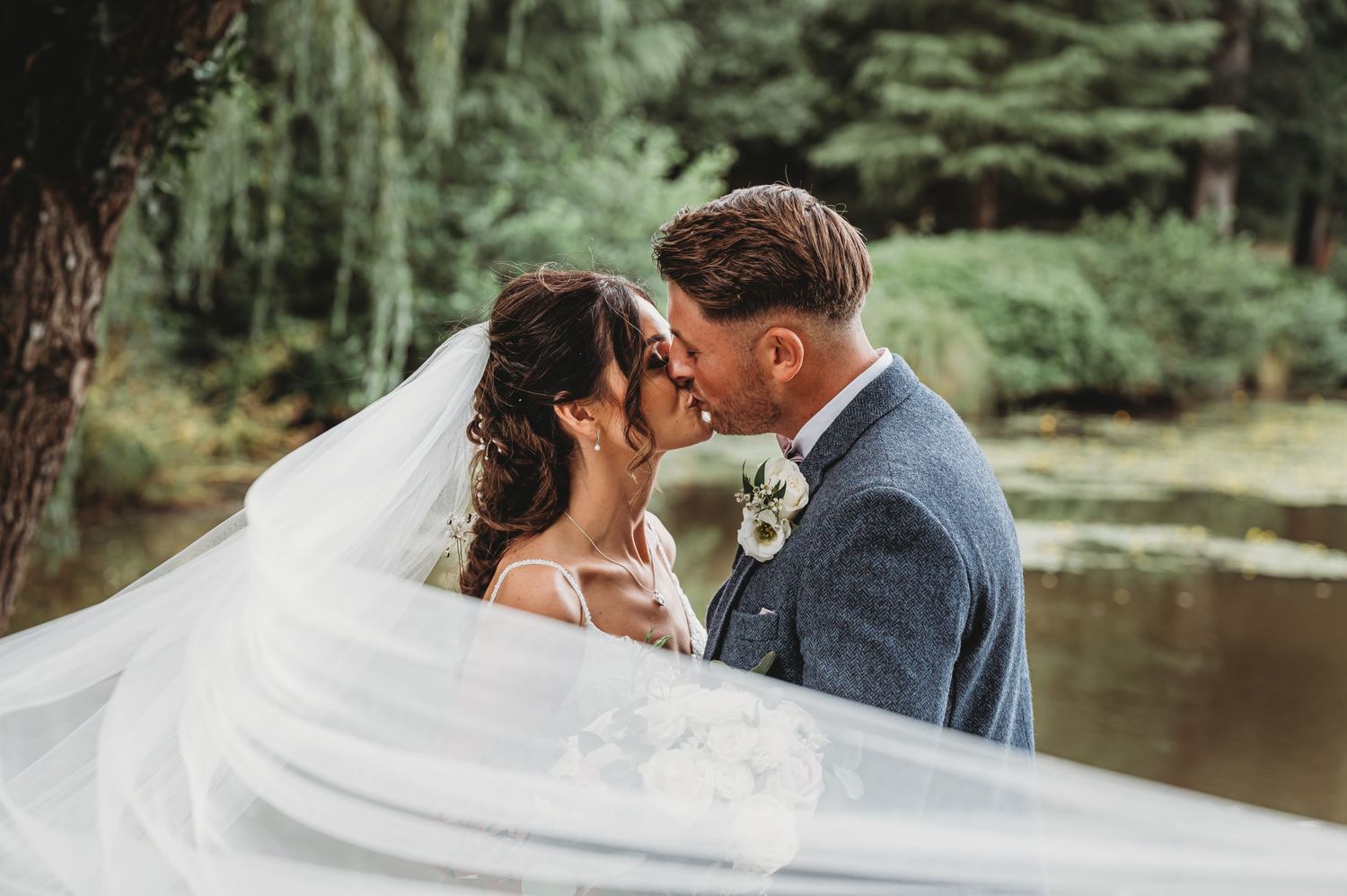 A romantic wedding couple shares an intimate moment as the bride's veil flows in the breeze by a peaceful pond.