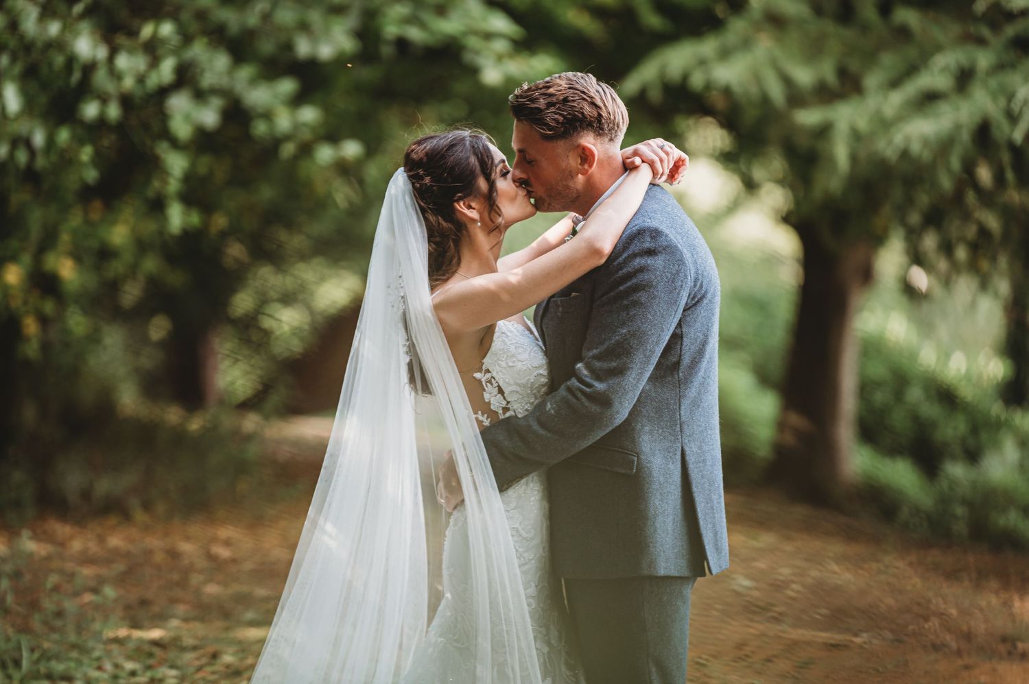 A romantic wedding scene between two people sharing a kiss on a tree-lined path with the bride wearing a flowing white veil.