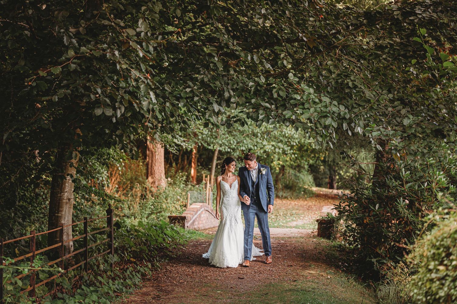 A couple stands together on a romantic tree-lined path in a lush garden setting during their wedding day.