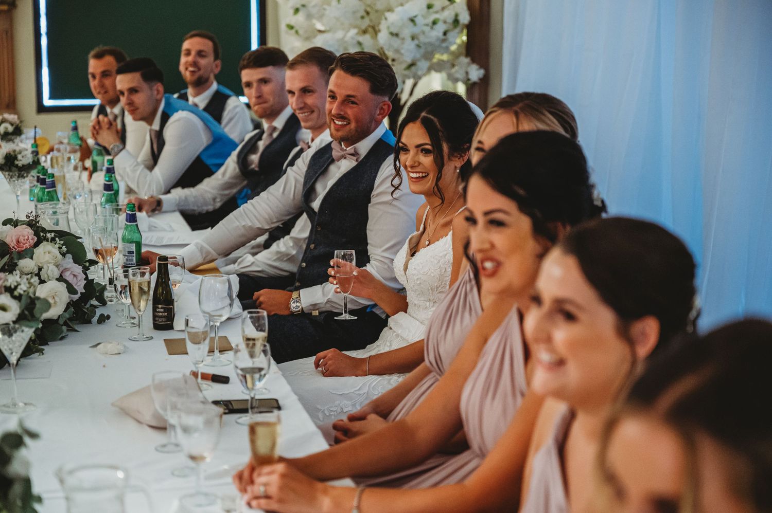 Wedding party guests in pink and gray attire seated at a long reception table with floral centerpieces.
