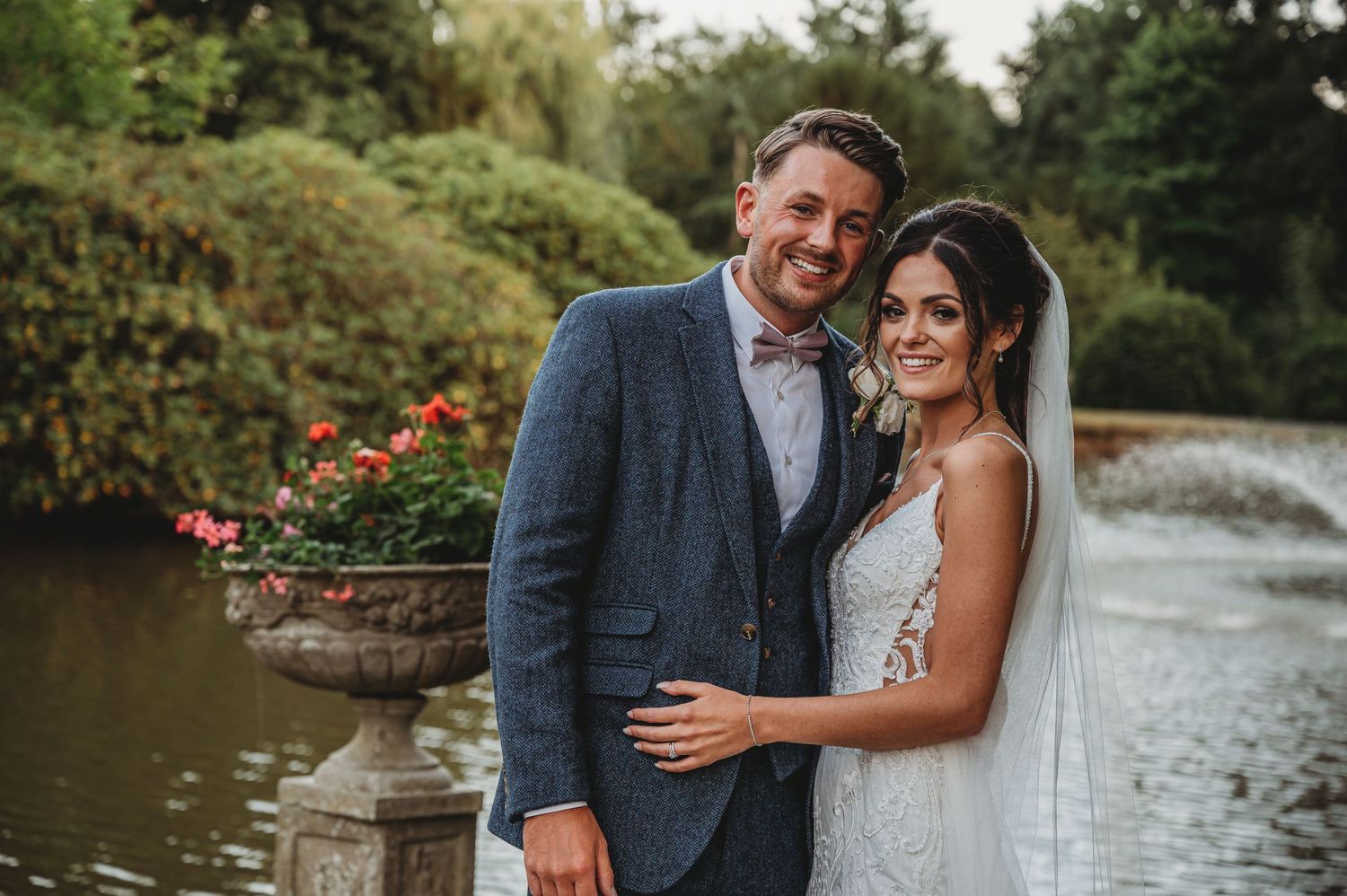 Couple in formal attire smiles together at outdoor garden venue with fountain in background at evening wedding.