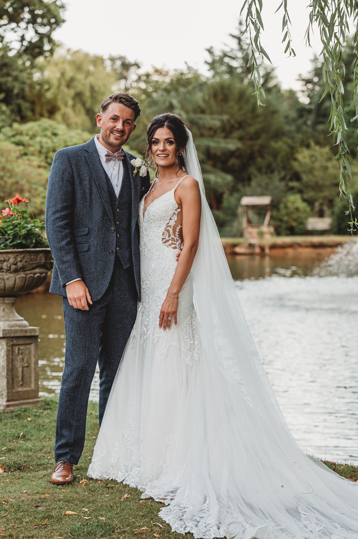 A newlywed couple poses in front of a garden pond, with the bride wearing a flowing lace gown and long veil.