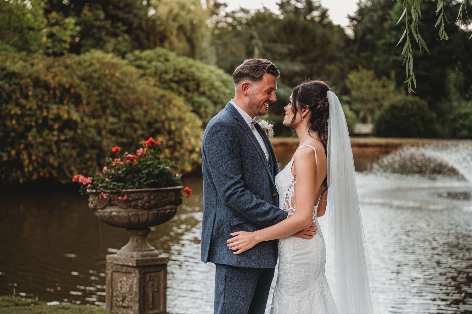 A romantic wedding couple embraces by a serene pond surrounded by lush gardens and blooming flowers.