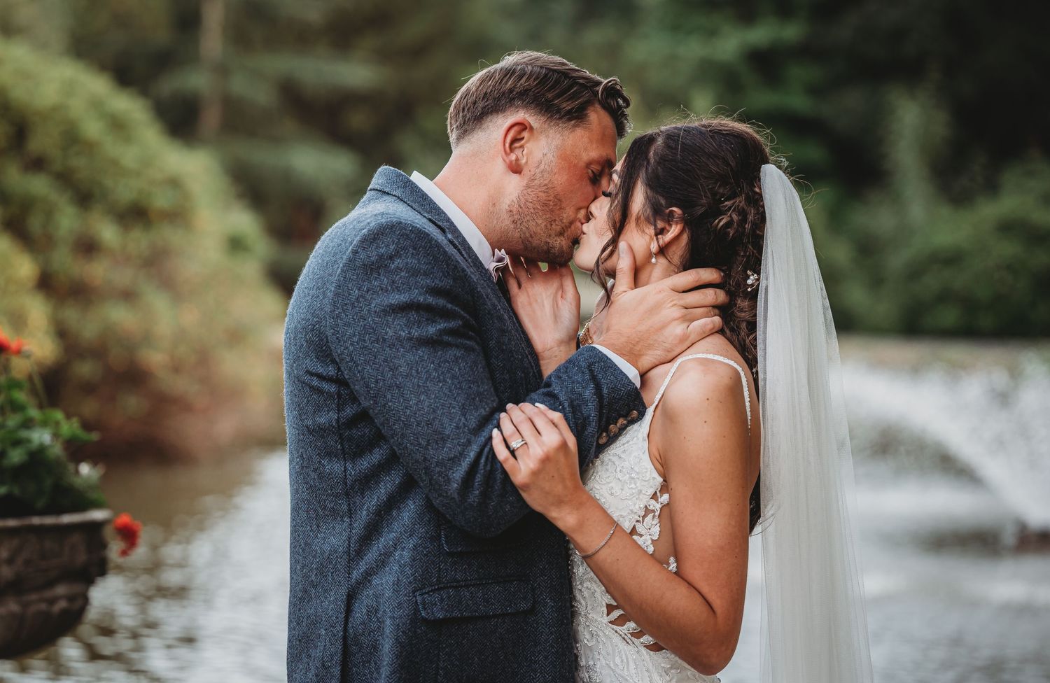 Newlyweds share a romantic kiss by a tranquil pond with trees and a stone bridge in the background.
