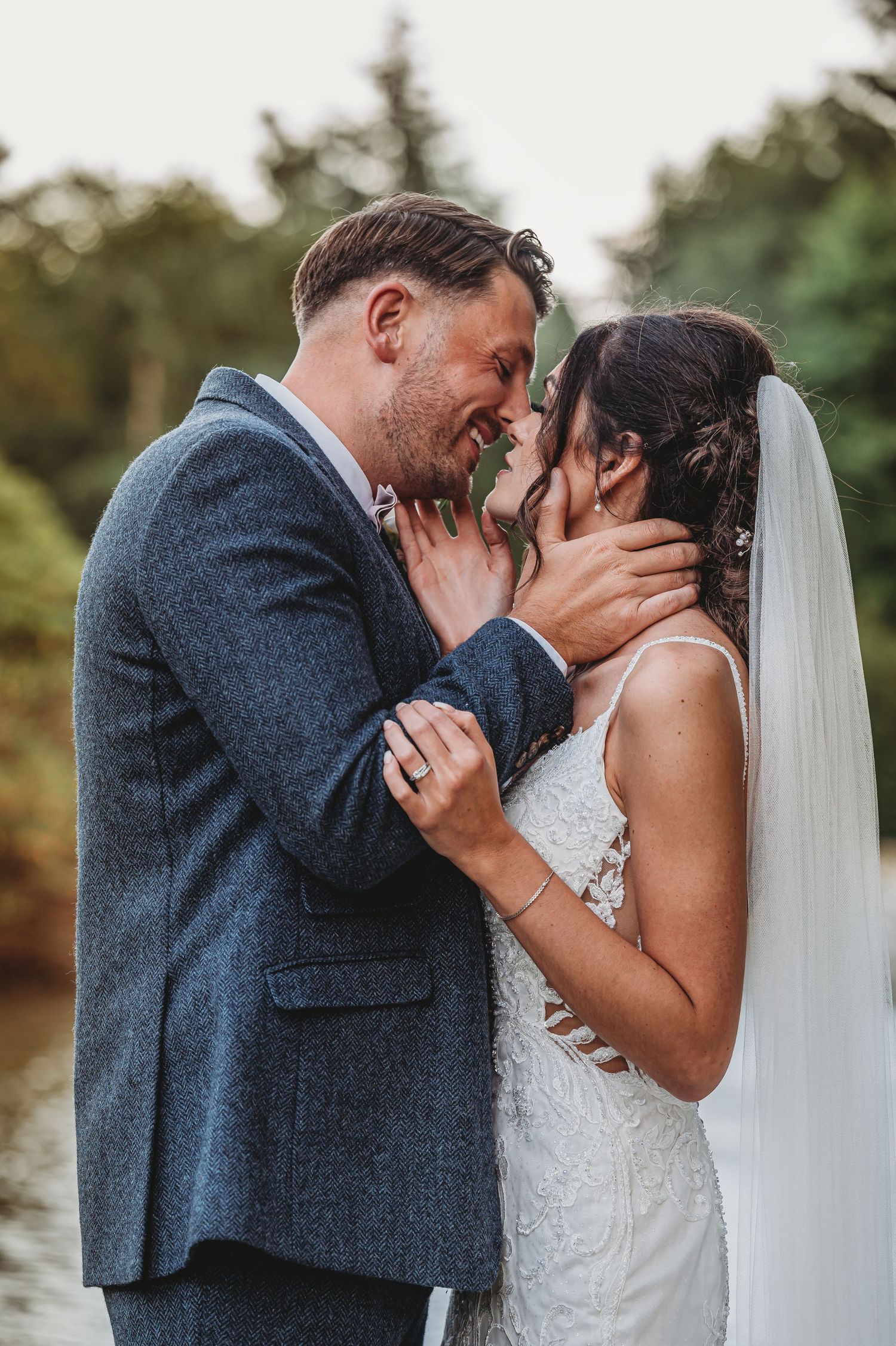 Newlywed couple shares intimate moment during outdoor wedding ceremony near trees and water.