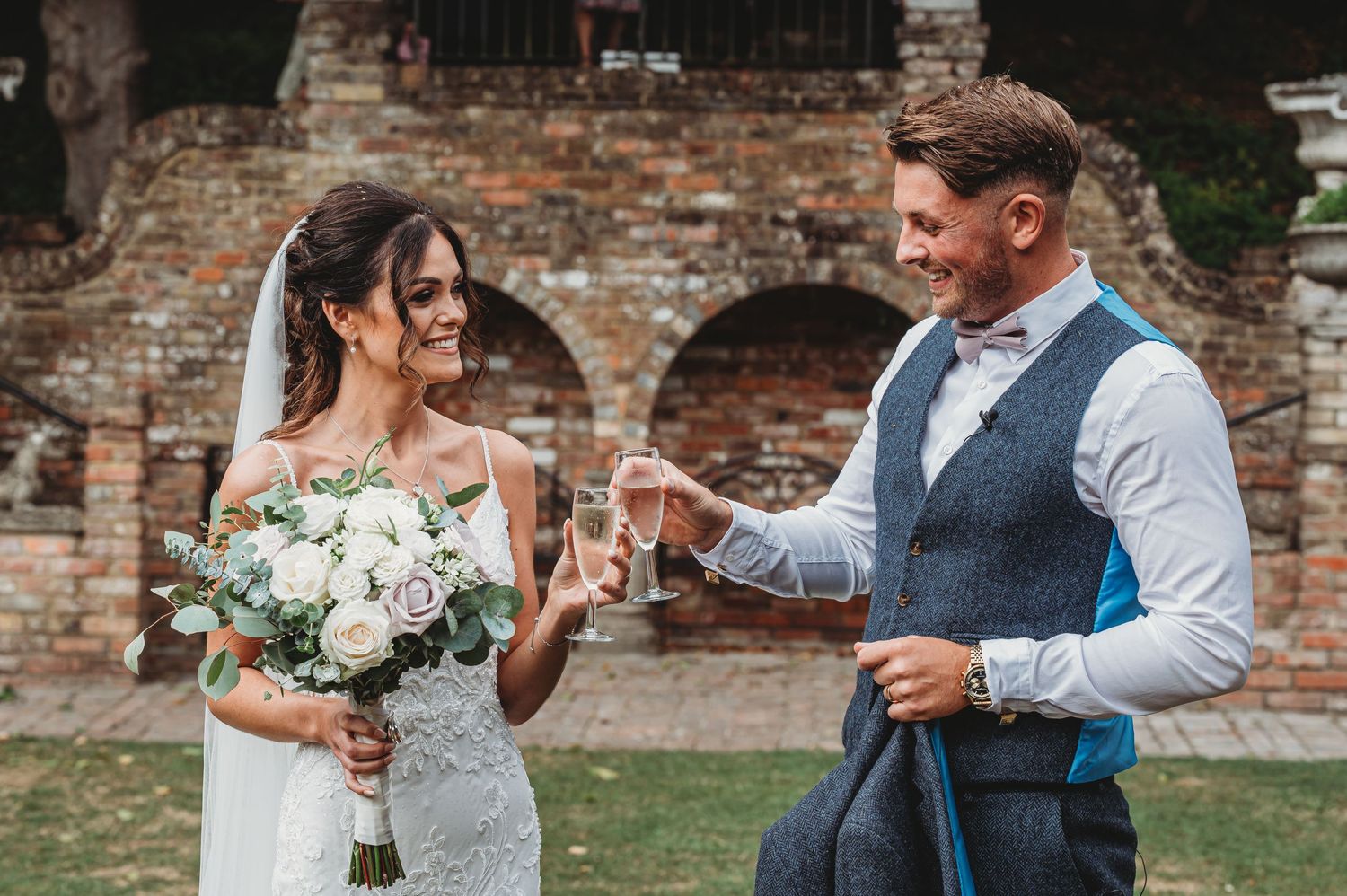 Happy couple toasts with champagne at rustic brick venue during wedding celebration with white floral bouquet.