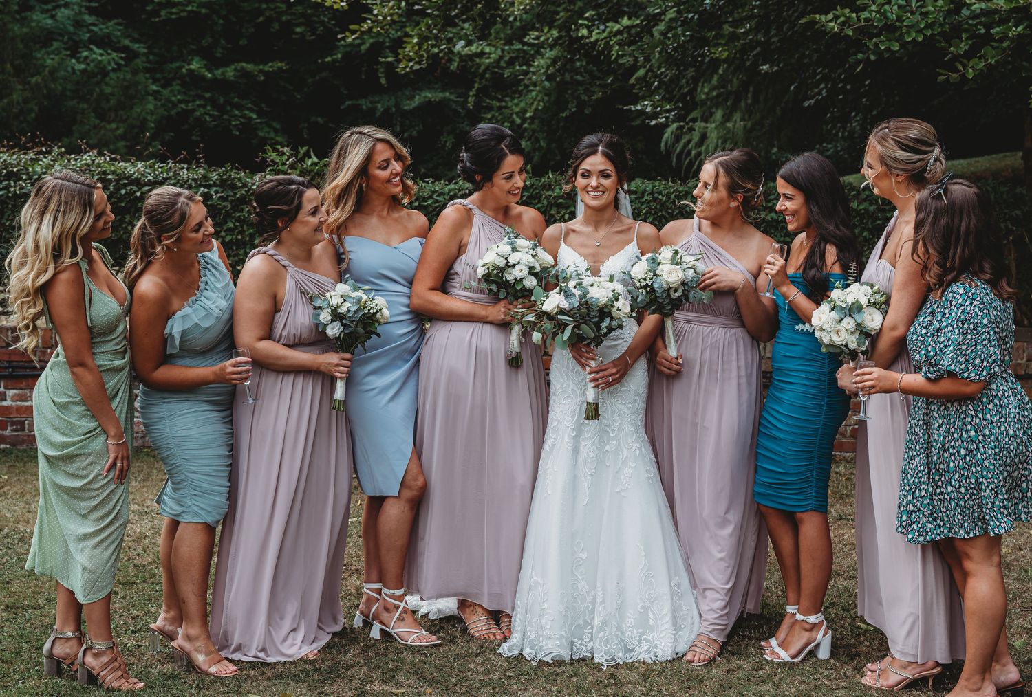 A group of bridesmaids wearing pastel colored dresses gather with the bride for a wedding photo in a wooded setting.