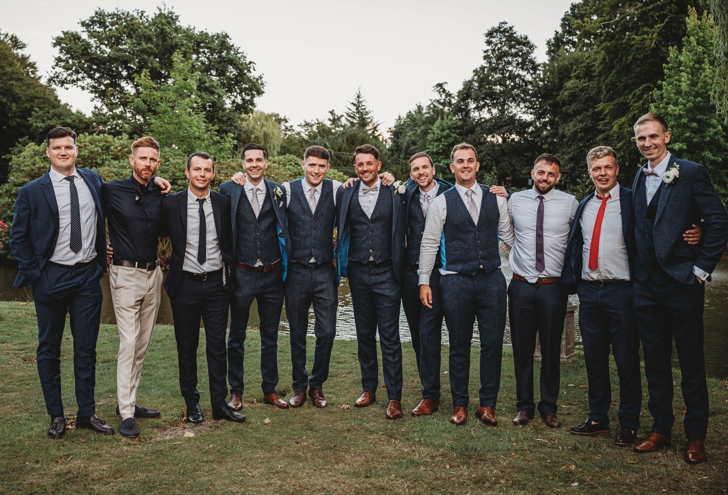 A group of well-dressed men stand together in formal attire on a grassy lawn with trees in the background.
