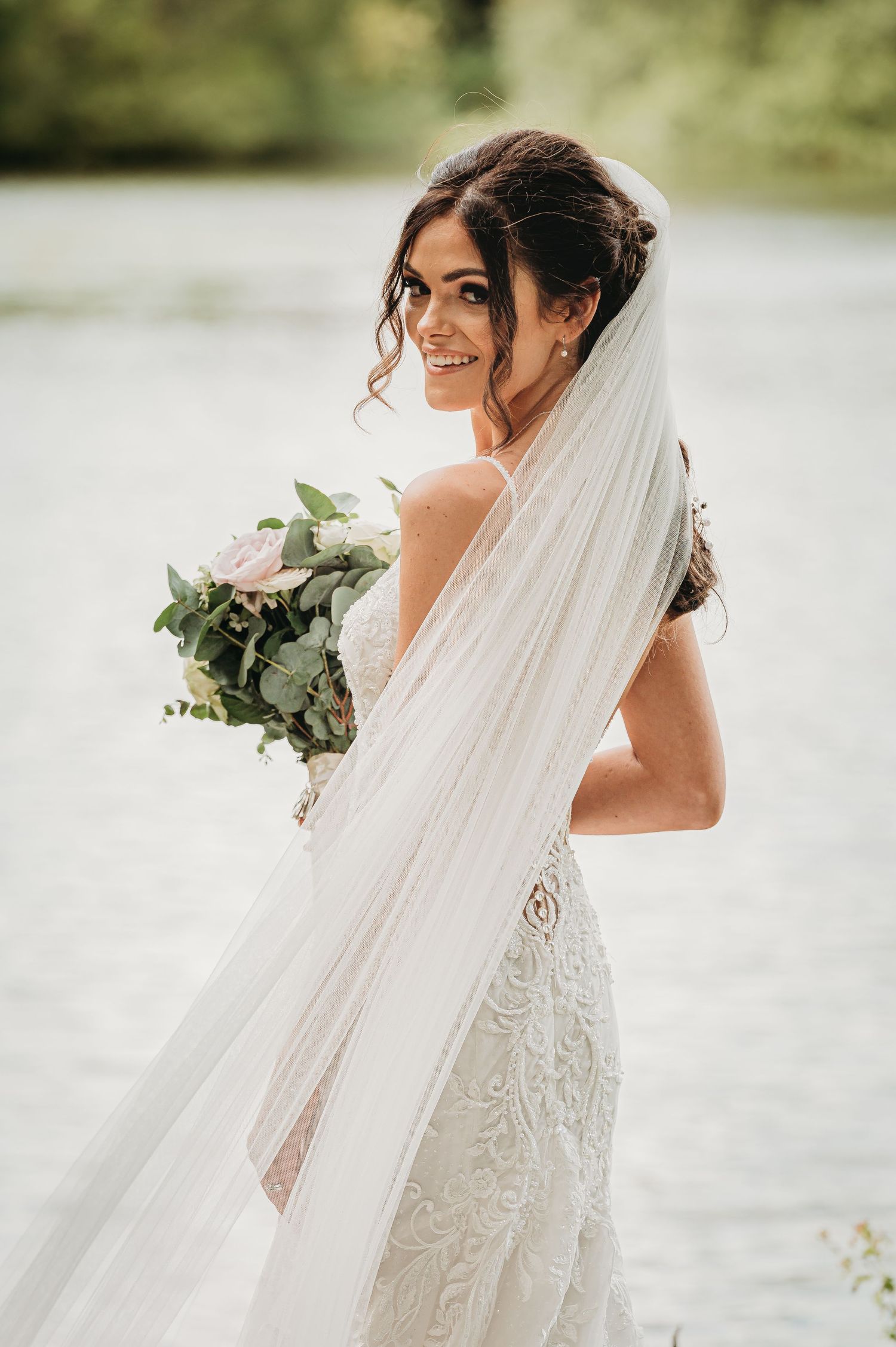 A bride in a lace wedding dress with flowing veil holds a white and green bouquet while posing by a lake.