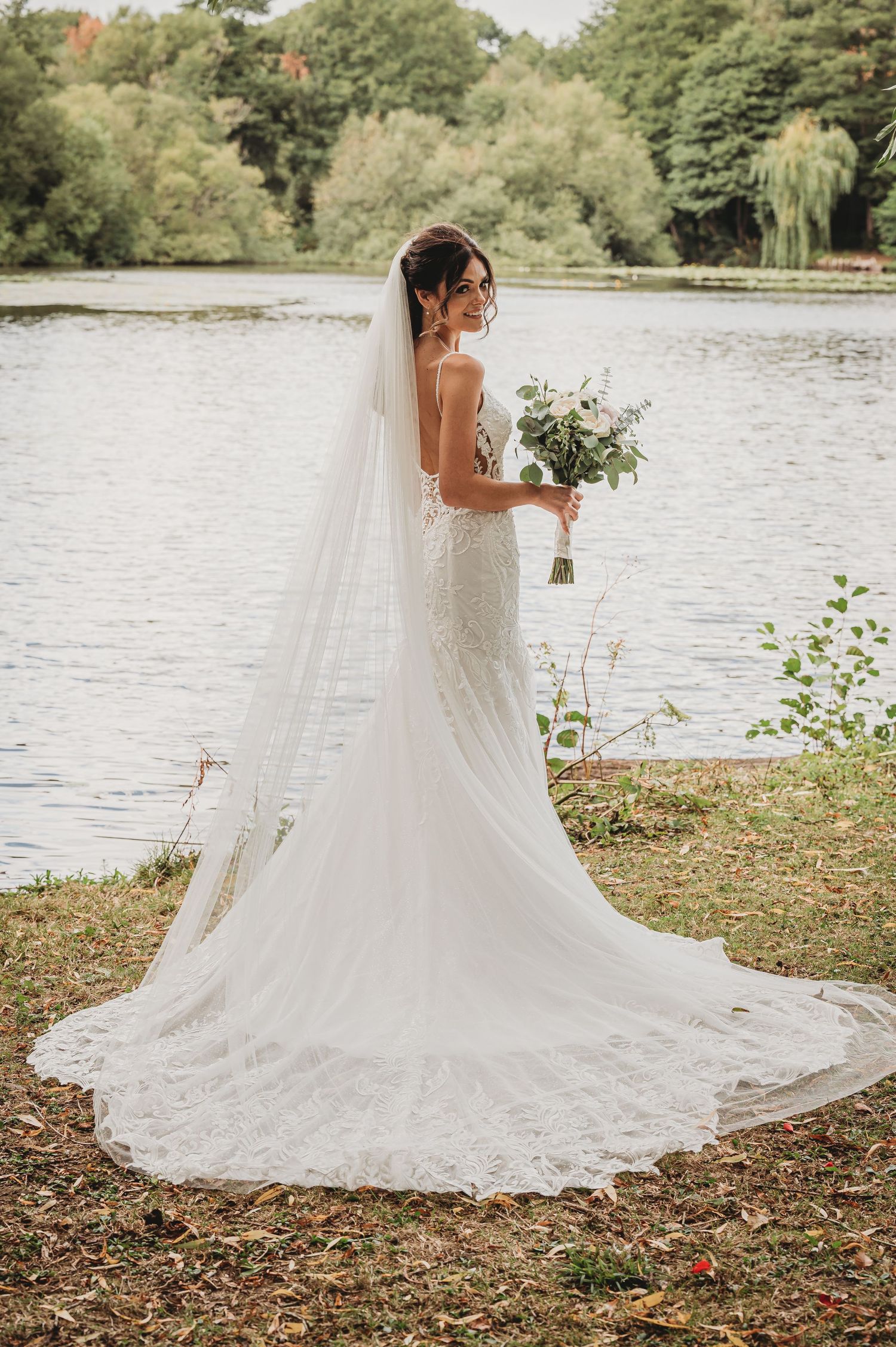 A bride in a lace wedding gown with long veil holds a bouquet beside a lake on her wedding day.