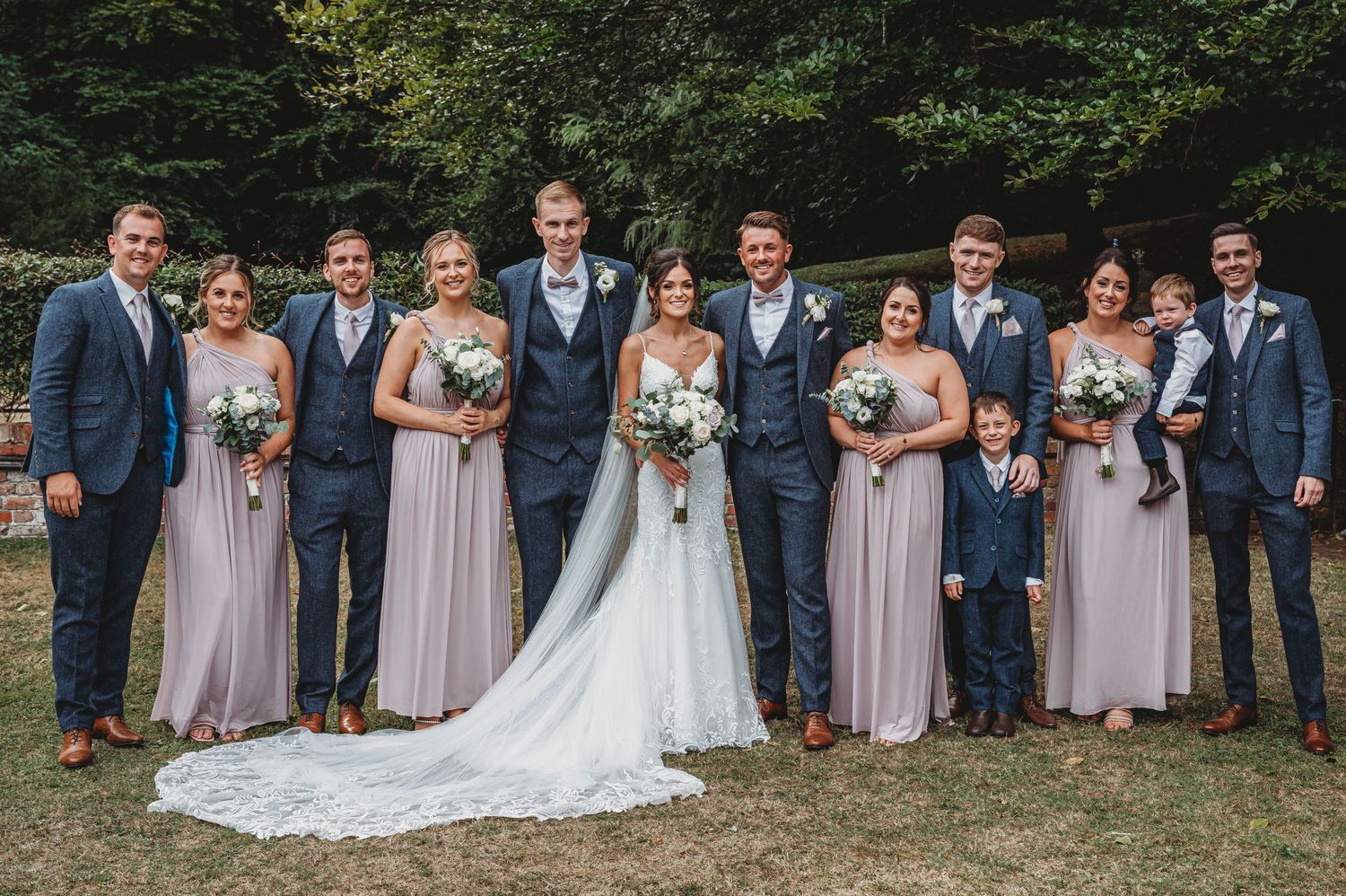 Wedding party wearing blue suits and pink bridesmaid dresses pose together outdoors in front of trees.