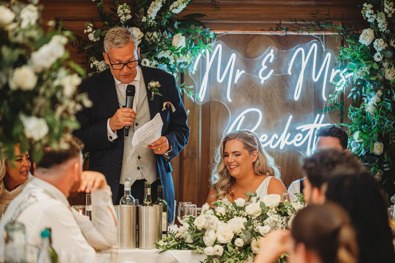 Wedding reception speech with neon Mr & Mrs sign glowing against floral backdrop.