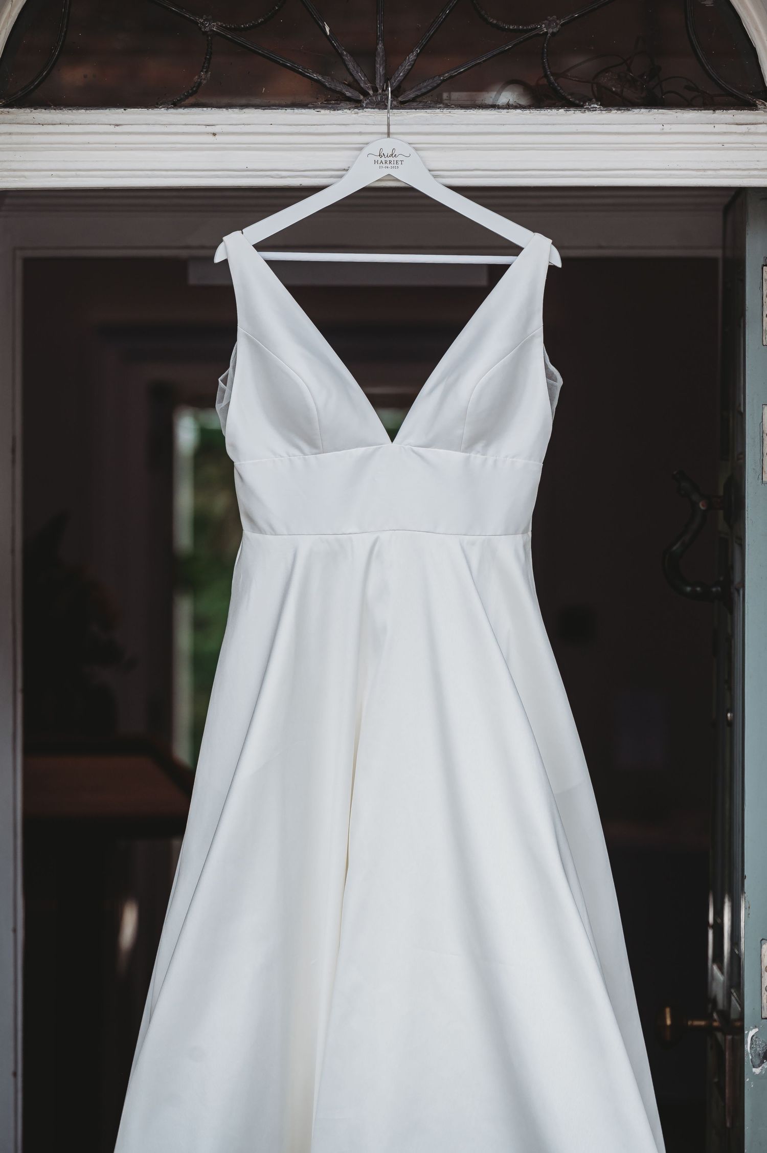 A classic white wedding dress with a plunging v-neckline and empire waist hangs on a white hanger against a dark background.