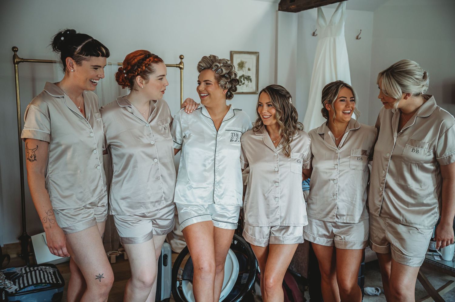 A group of friends laugh and smile together while wearing matching light gray satin pajama sets in a bedroom setting.