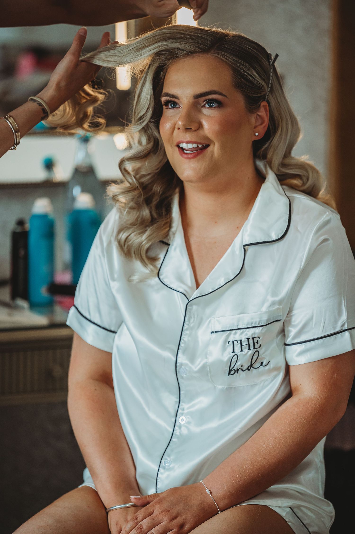 A smiling individual wearing a white monogrammed pajama shirt with "THE bride" text sits during wedding preparation.