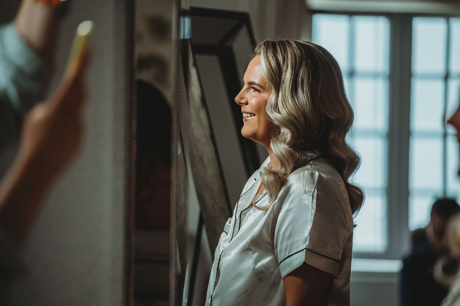Someone with wavy blonde hair stands in profile wearing a light colored button-up shirt in an indoor setting.