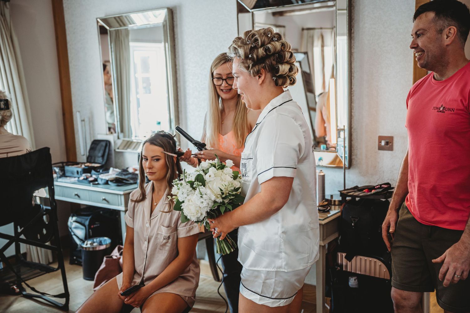 People sharing a joyful moment during wedding preparations at a salon with hair curlers and bouquet.