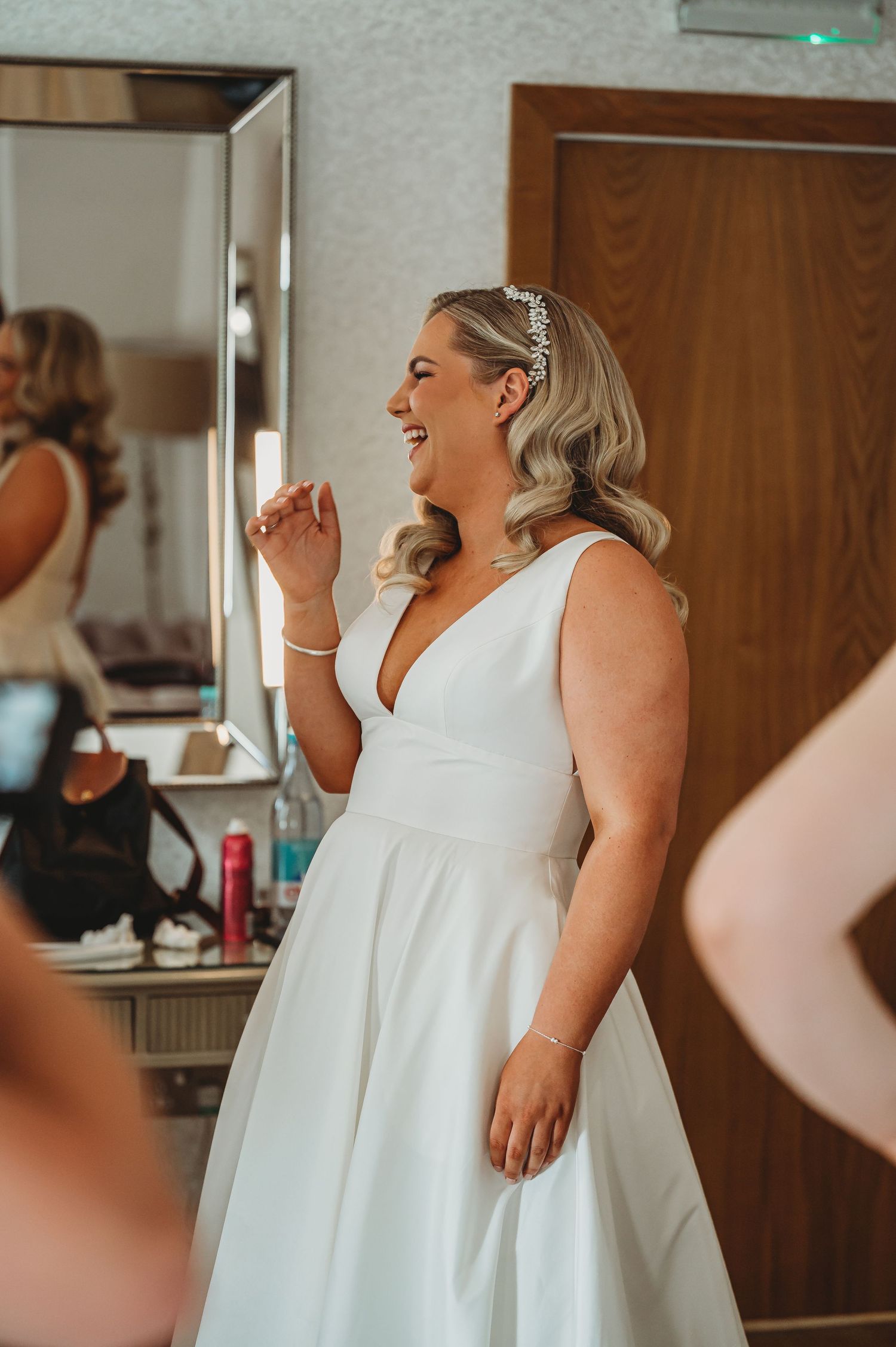 A bride wearing an elegant white V-neck wedding dress smiles joyfully while getting ready in a dressing room.